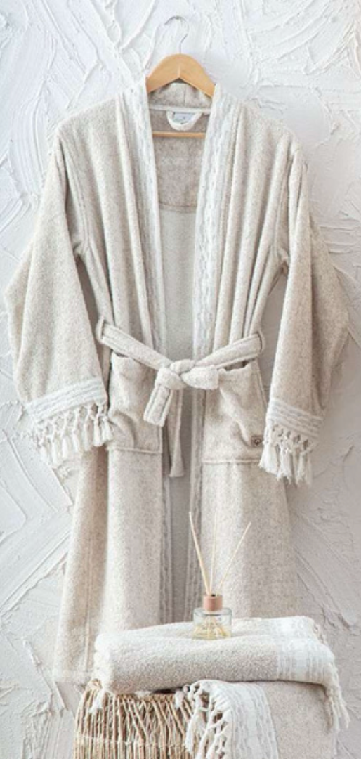 Beige textured bathrobe with tassel trim hanging against a white wall, folded towel and diffuser in foreground — luxury eco-conscious home textile style.