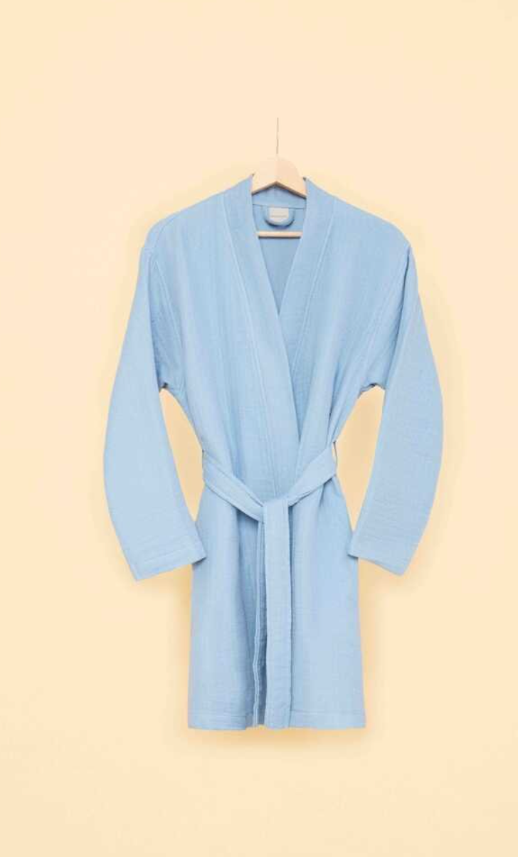 Sky blue bathrobe with waist tie hanging on wooden hanger against cream-colored wall — modern sustainable homewear with minimalist design.