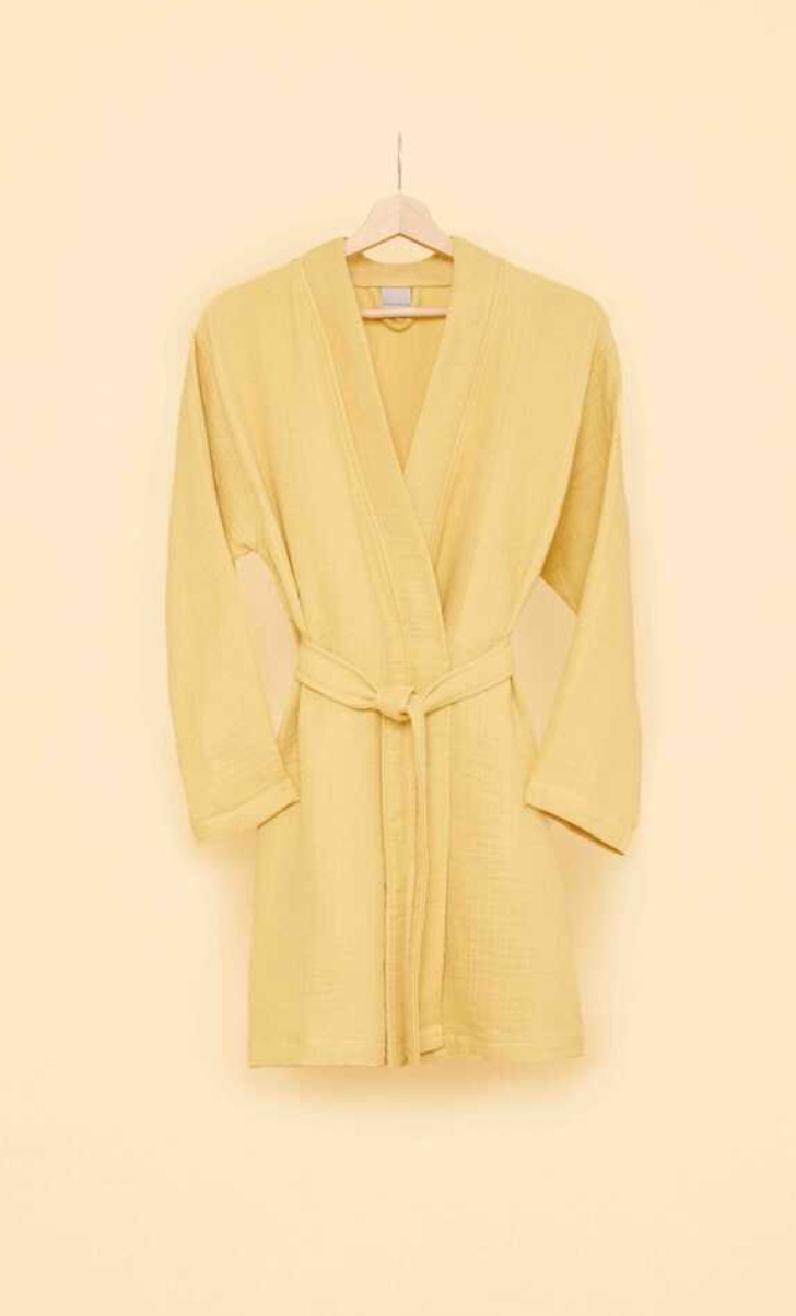 Soft yellow bathrobe with belt tie on wooden hanger against a warm beige wall — trendy eco-conscious loungewear for sustainable living.
