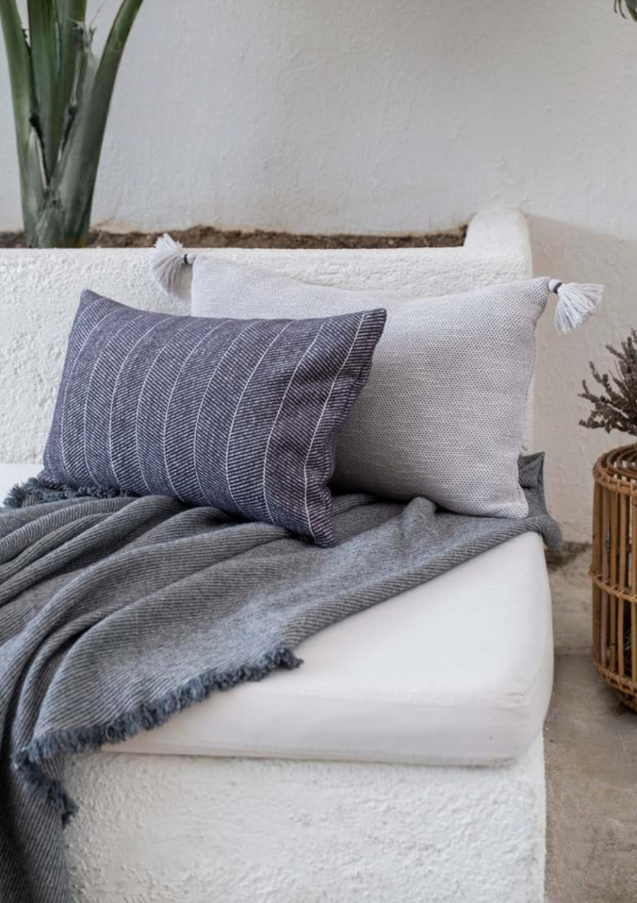 Bone-colored and navy striped pillows with tasseled corners styled on a textured sofa with a grey fringed throw — modern eco-luxury home decor.